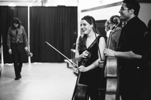 Lauren is backstage, laughing without restraint while holding her cello, wearing an evening gown. Kevin is next to her, blurred, but clearly also laughing. John Whaley is blurred in the far background looking pensive