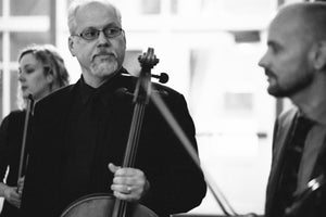 Collin is backstage holding his cello, looking fairly serious, but optimistic maybe? Valdine is blurred behind him, and Doug is blurred in the foreground.