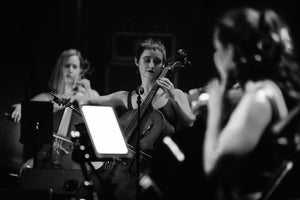 Sage is looking quite glamorous on stage, about to make a long upbow, her eyes are closed, and there's some glitter evident from her make-up. Other cellists are blurred in the background and foreground.